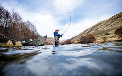 Angler steelhead fly fishingon the Deschutes river with a drift boat, mountains and trees in the background