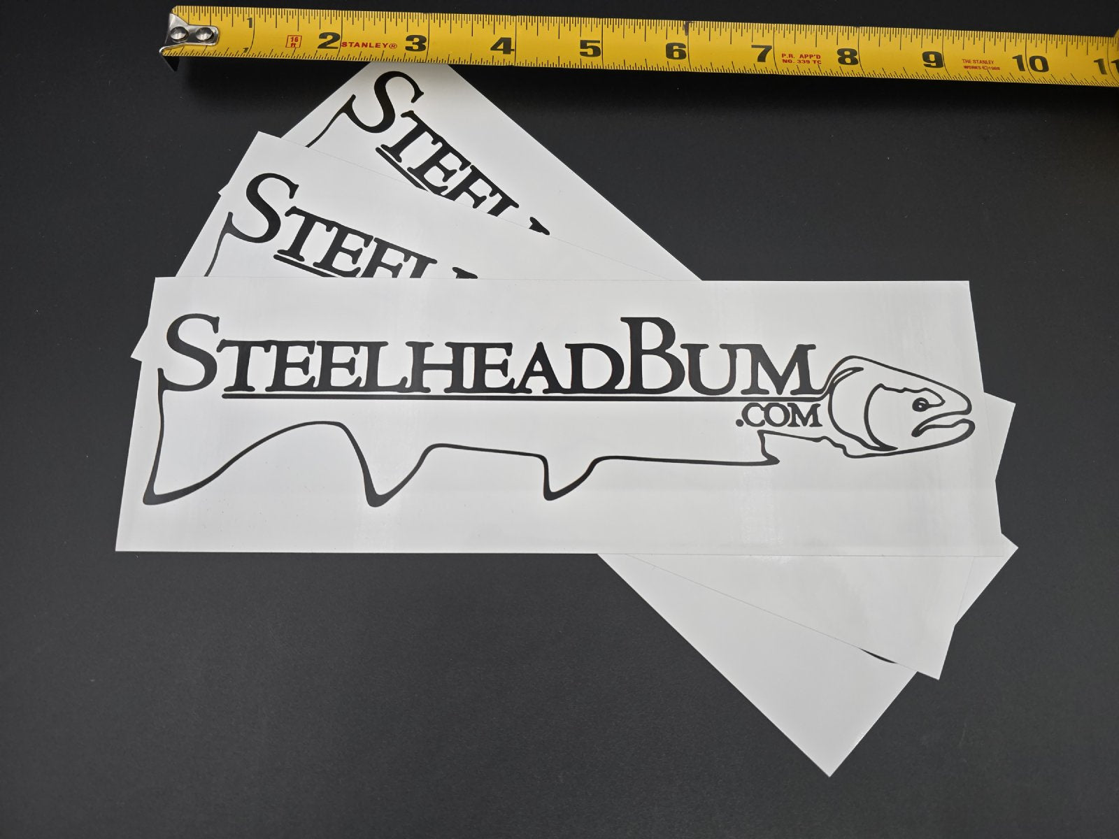 Three Large 'SteelheadBum.com' stickers with a measuring tape at the top on a dark background