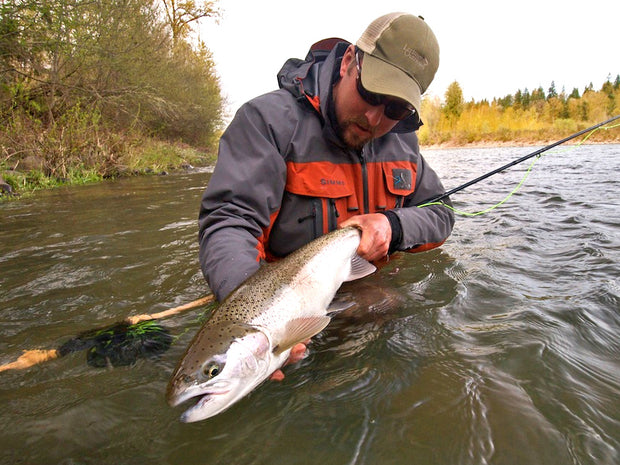 Tom Larimer fishing for Steelhead, holding a caught fish while in a river with trees and foliage in the background